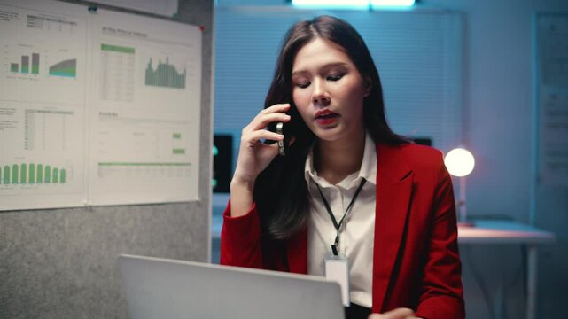 Young Asian manager wearing red jacket working late at night in office using laptop and talking on phone