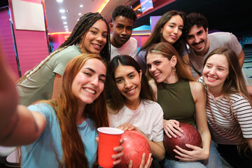 Cheerful young friends are posing for a selfie while holding bowling balls and drinks at the bowling alley