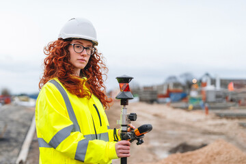 Portrait of confident female land surveyor with long hair in bright safety gear using surveying...