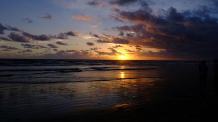 A breathtaking sunset view at a beach in Bali, Indonesia, capturing the golden sun dipping into the ocean, dramatic clouds, and warm reflections on the wet shoreline.