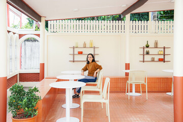 Joyful brunette curly woman in trendy pants and shirt smiles and looks out to view Pretty lady in jeans sits on chair in cafe