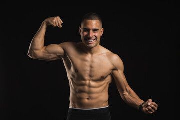 Fototapeta premium Smiling muscular man flexing biceps and abs in front of black background. Lifestyle. Sport.