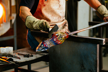 A glassblower shapes colorful molten glass using tools in a workshop.