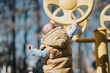 A small child in a hooded jacket interacts with playground equipment on a sunny day, showcasing...