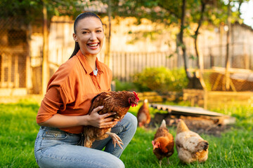 Portrait of happy young woman on private farm holding chicken and smiling at camera, posing on backyard farm, promoting organic poultry farming © Home-stock