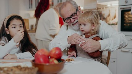 A grandfather sits at a table and wipes the hands of his grandson who is sitting on him with a napkin, while his granddaughter sits next to them and draws with pencils - Powered by Adobe