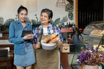 Two Asian female bakery owners smiling while standing in front of glass display, one holding bread basket with tongs, another using tablet. Representing teamwork, digital operations and local cafe 