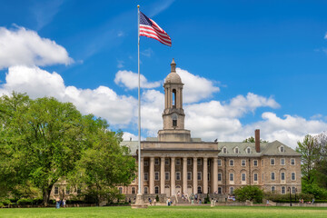 The Old Main building on the campus of Penn State University and American flag in spring sunny day, State College, Pennsylvania.