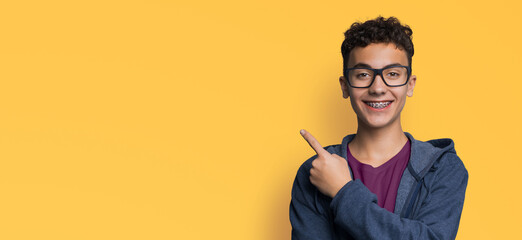 Excited happy black curly hair man in braces brackets, glasses, hoodie jaket, purple t-shirt advertise show point sale slogan text area, isolate yellow background. Dental care, ophthalmology ad