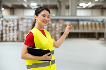 Smiling woman in high visibility vest giving directions inside a warehouse while holding a digital...