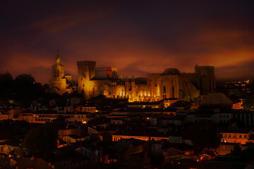 Golden lights bathe the medieval Palais des Papes in Avignon, France, under a moody twilight sky....