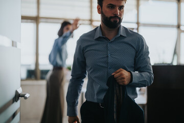 A focused businessman leaves a meeting room, holding a jacket, as a colleague gestures in the background. The scene conveys determination and professionalism in a modern office setting.