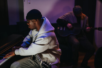 A focused musician plays the piano while another strums a guitar in a cozy studio. The dim lighting...