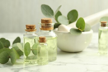 Bottles of essential oil, eucalyptus leaves, mortar and pestle on white marble table, closeup