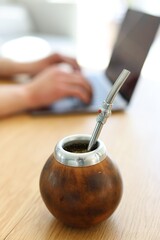 Man using laptop at wooden table indoors, focus on traditional yerba mate tea