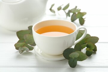 Aromatic tea in cup and eucalyptus branches on white wooden table, closeup