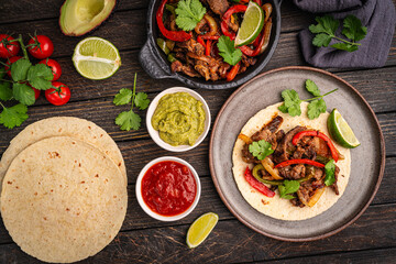 Fajitas, beef meat slices with colored pepper and onions, traditional mexican food, served with tortillas, salsa and sour cream on wooden background, top view. High quality photo