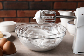 Hand mixer with whisks and whipped cream on wooden table against brick wall, closeup