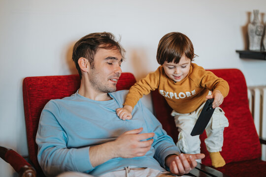 A father and his toddler son bonding while using a smart phone on a cozy red chair, creating a warm and playful family atmosphere. - Powered by Adobe
