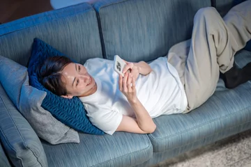  ソファに寝転んでスマホを使う女性 © naka