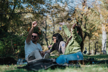 Three friends having fun outdoors, tossing playing cards into the air. The scene captures joy and friendship on a sunny day in a natural setting.