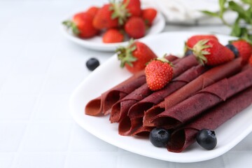 Tasty fruit leather rolls, strawberries and blueberries on white table, closeup. Space for text