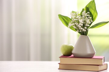 Beautiful lilies of the valley flowers in vase, apple and books on white table against blurred background. Space for text