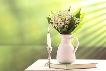 Beautiful lilies of the valley flowers in vase, candle and book on white table against blurred...