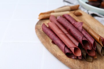 Tasty fruit leather rolls on white tiled table, closeup. Space for text