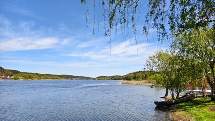 A wooden boat is moored under the trees on the grassy shore of the lake. There are houses on the far shore. A mixed forest grows around the lake. On the water ripples Sunny spring weather and blue sky
