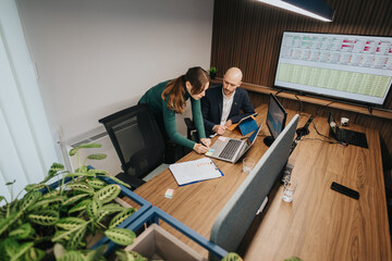 Two associates discuss and evaluate business strategies at a desk using laptops and charts,...