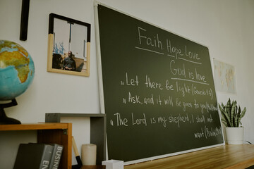 Blackboard displaying inspirational religious quotes with bookshelf and globe on desk nearby. Photo conveying an atmosphere of faith and knowledge in study or classroom setting