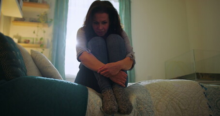 Woman sitting on bed, tightly clutching knees, crying in anguish, themes of despair, emotional...