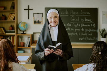 Fototapeta premium Portrait of cheerful nun standing in classroom holding open book teaching students sitting at desks during lesson on faith and religion educational setting