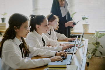 Students typing on laptops while being supervised by teacher. Classroom setting with natural elements like plants and light from windows providing calm environment
