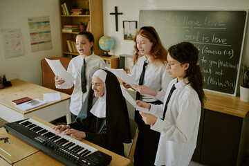 Obraz premium Elderly nun playing electric piano while students sing in classroom decorated with educational materials and cross on wall showing group engaged in musical activity