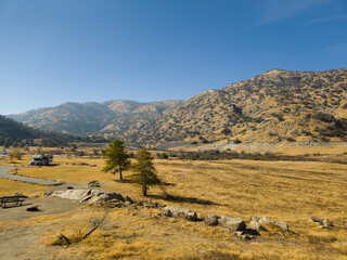 A camper van parked in a dry, open desert valley surrounded by rugged mountains in California. The golden-brown landscape under a clear blue sky.