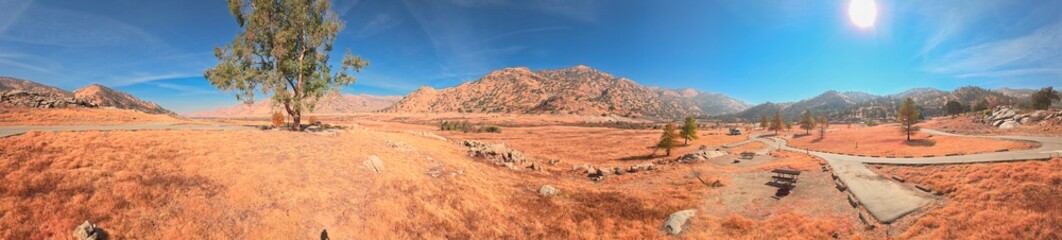 Panoramic view of a dry, sunlit mountain valley in California, featuring golden-red grass, winding paved paths, and scattered trees under a clear blue sky.