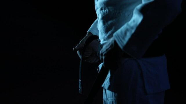 Close-up: Martial artist in gi meticulously ties their black belt in slow motion. Dark, moody atmosphere with cool blue lighting emphasizes focus and pre-fight ritual.