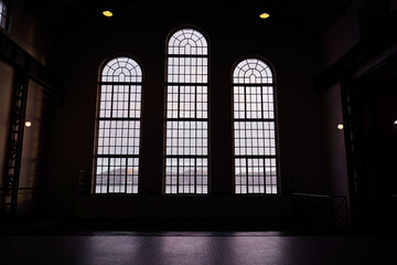 Three large arched windows with grid patterns, casting soft light into a dimly lit interior space. The view outside is obscured, creating a moody atmosphere.