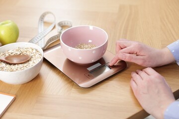 Woman weighting oatmeal on kitchen scale at wooden table, closeup