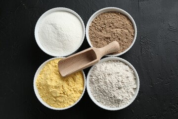 Different types of flour in bowls and scoop on black textured table, top view