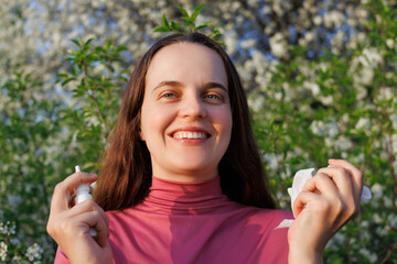 Woman smiles, relieved from seasonal allergies, holding nasal spray tissue outdoors amidst...