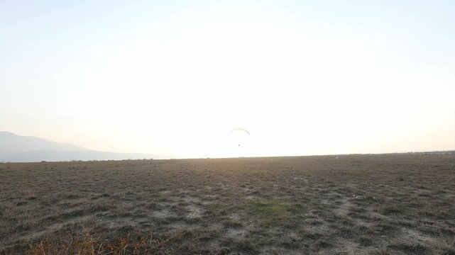 Lone powered paraglider gracefully flies across a hazy, atmospheric sky during sunset. Below, distant silhouetted hills create a serene, minimalist backdrop for this adventure sport.