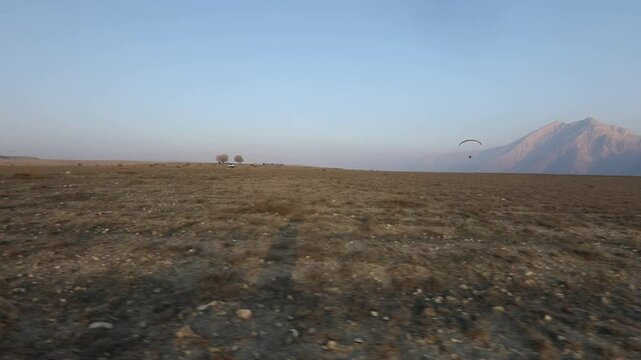 Lone powered paraglider gracefully flies across a hazy, atmospheric sky during sunset. Below, distant silhouetted hills create a serene, minimalist backdrop for this adventure sport.