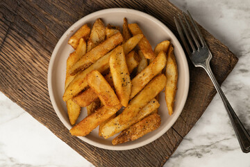 Crispy, golden, seasoned, homestyle french fries on a plate with a fork on wood cutting board and marbled background.