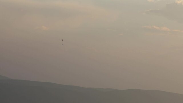 Lone powered paraglider gracefully flies across a hazy, atmospheric sky during sunset. Below, distant silhouetted hills create a serene, minimalist backdrop for this adventure sport.