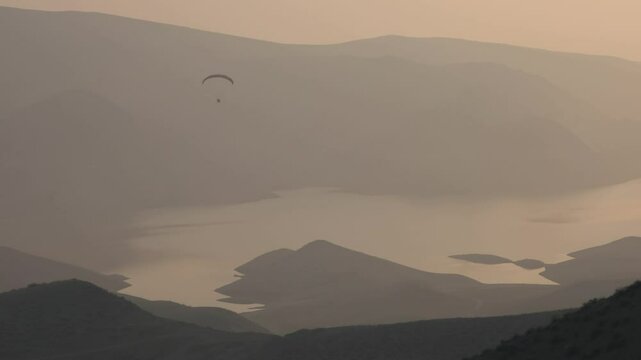 Lone powered paraglider gracefully flies across a hazy, atmospheric sky during sunset. Below, distant silhouetted hills create a serene, minimalist backdrop for this adventure sport.