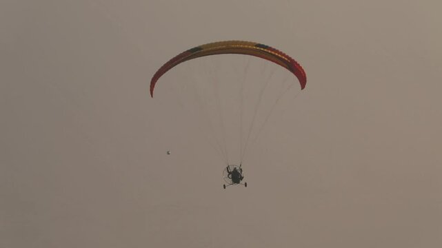Lone powered paraglider gracefully flies across a hazy, atmospheric sky during sunset. Below, distant silhouetted hills create a serene, minimalist backdrop for this adventure sport.