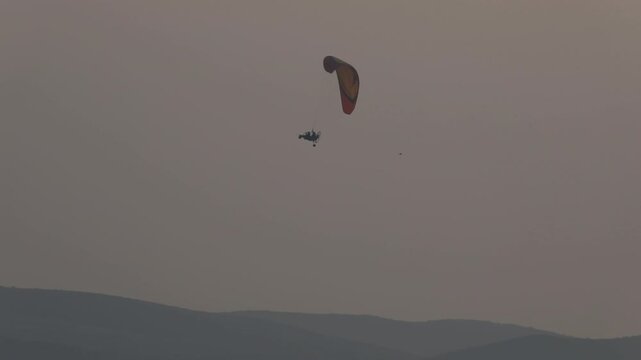 Lone powered paraglider gracefully flies across a hazy, atmospheric sky during sunset. Below, distant silhouetted hills create a serene, minimalist backdrop for this adventure sport.